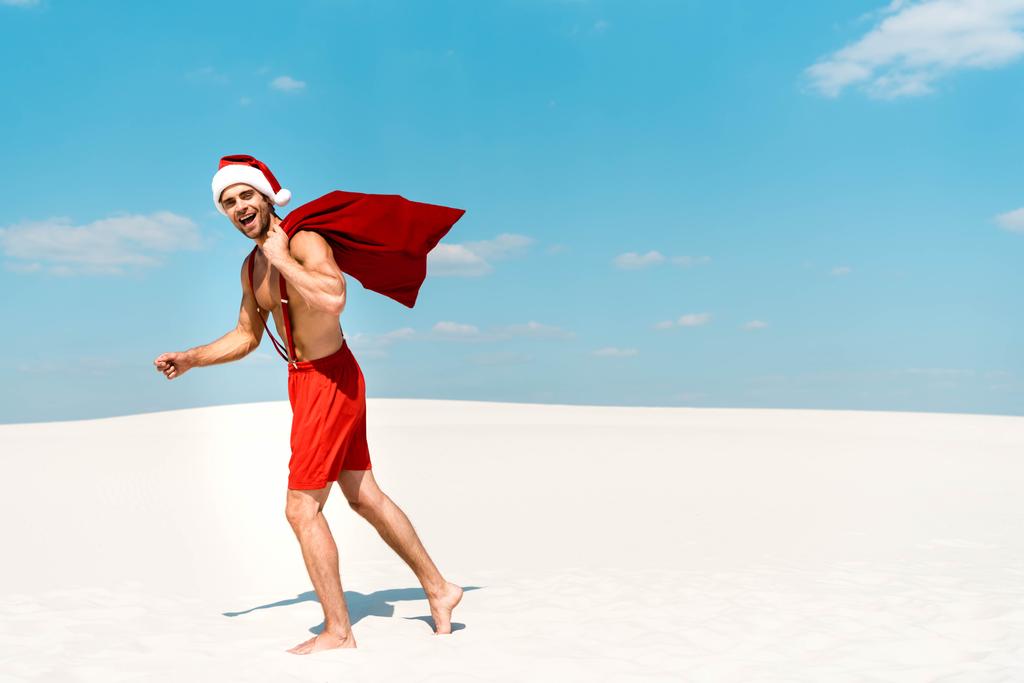 όμορφος και σέξι άντρας με καπέλο santa κρατώντας σάκο santa και περπατώντας στην παραλία στις Μαλδίβες  - Φωτογραφία, εικόνα