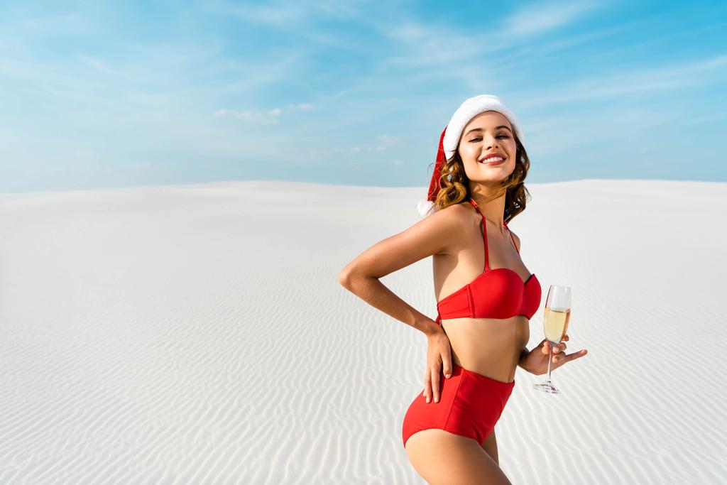 sexy e sorridente donna in cappello di Babbo Natale e costume da bagno in possesso di bicchiere di champagne sulla spiaggia in Maldive
  - Foto, immagini
