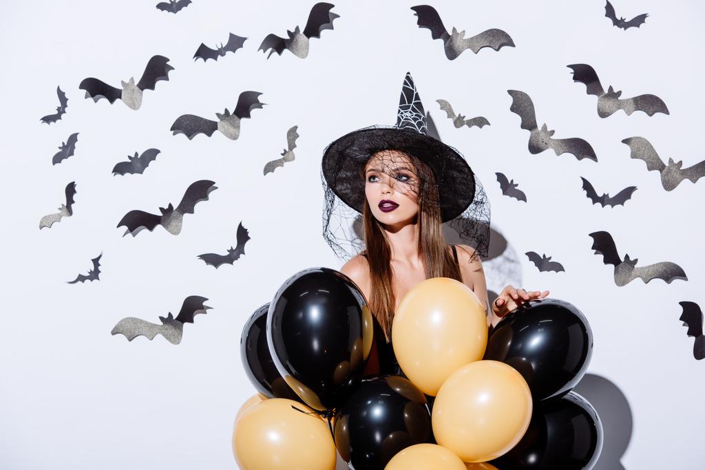 dívka v černé čarodějnice Halloween kostým drží balónky v blízkosti bílé zdi s dekorativními netopýry - Fotografie, Obrázek