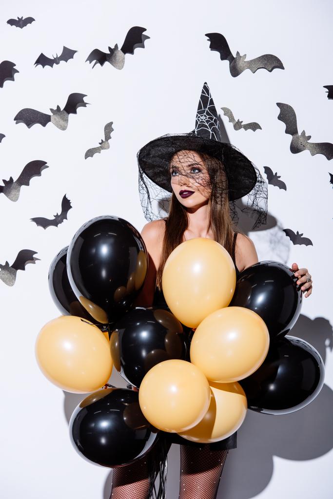 ragazza in costume strega nera Halloween tenendo palloncini vicino muro bianco con pipistrelli decorativi
 - Foto, immagini