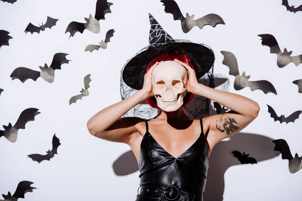 ragazza in costume nero strega Halloween con i capelli rossi tenendo teschio davanti al viso vicino muro bianco con pipistrelli decorativi
 - Foto, immagini