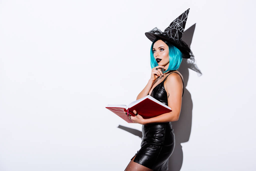 nachdenklich sexy Mädchen im schwarzen Hexenhalloween-Kostüm mit blauem Haar, das Buch auf weißem Hintergrund hält - Foto, Bild