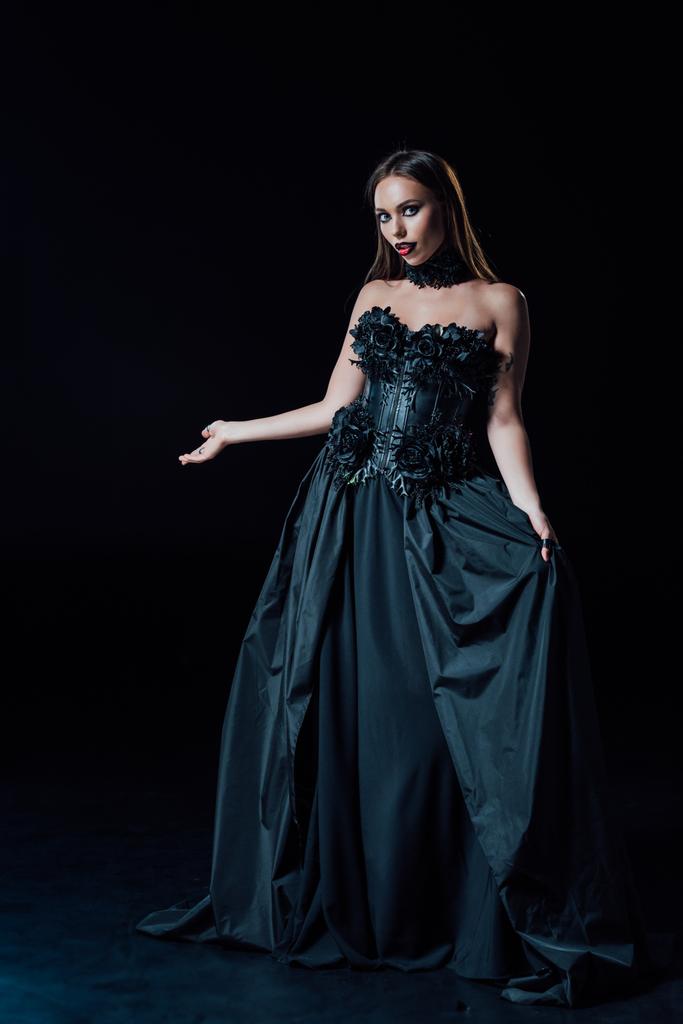 asustadiza chica vampiro en vestido gótico negro apuntando con la mano sobre fondo negro
 - Foto, Imagen