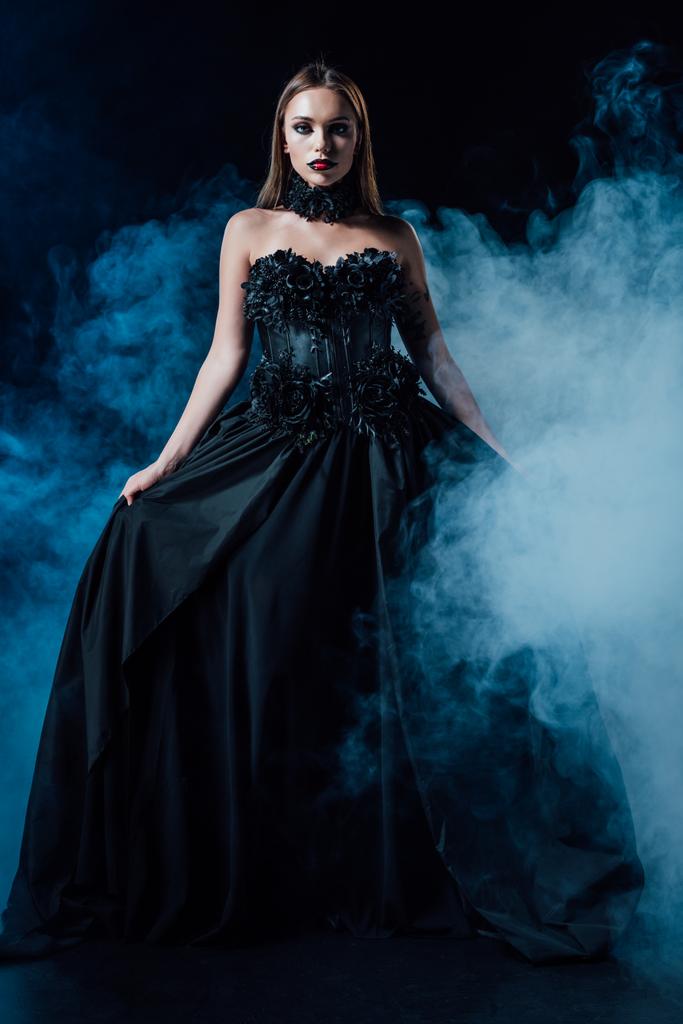 τρομακτικό κορίτσι βαμπίρ σε μαύρο γοτθικό φόρεμα σε μαύρο φόντο με καπνό - Φωτογραφία, εικόνα