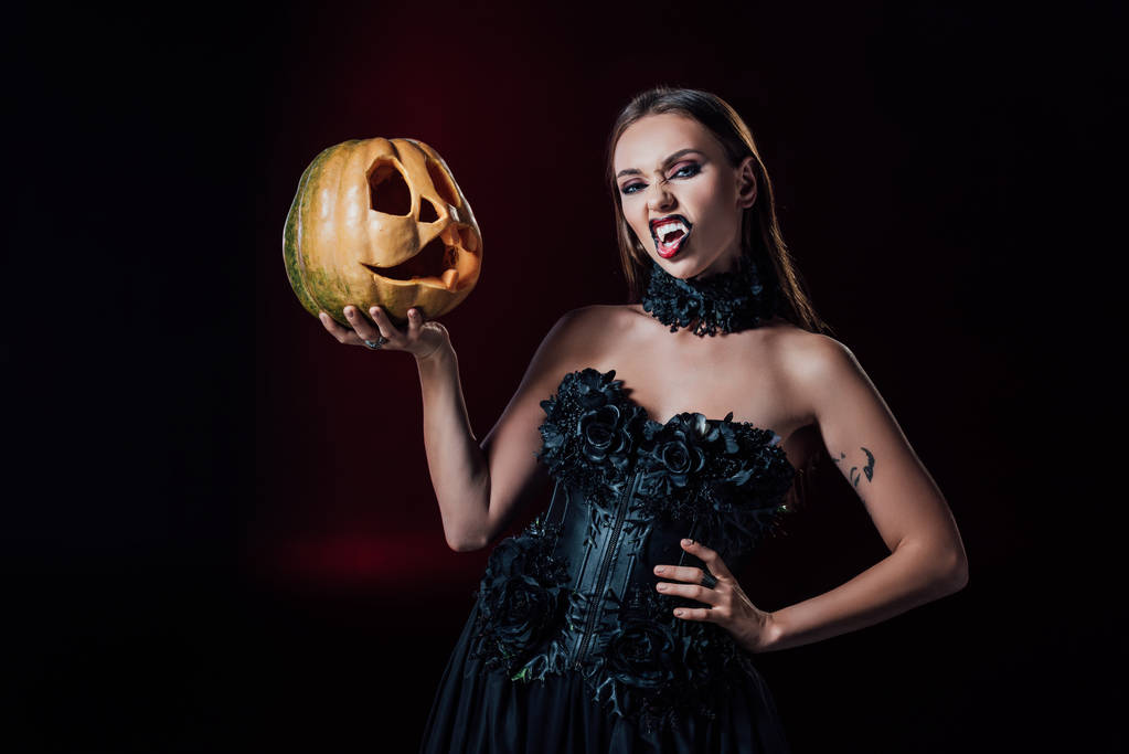 scary vampire girl with fangs in black gothic dress holding Halloween pumpkin on black background - Photo, Image