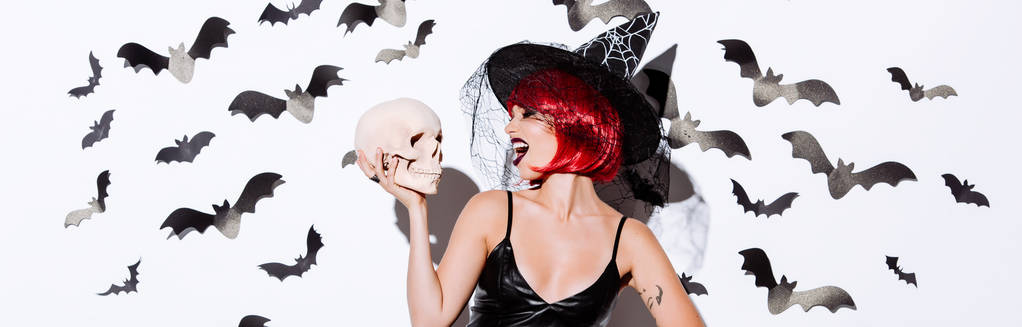 Панорамный снимок девушки в костюме черной ведьмы на Хэллоуин с рыжими волосами, держащими череп возле белой стены с декоративными летучими мышами
 - Фото, изображение