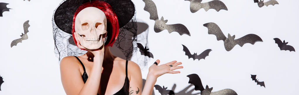 Panoramaaufnahme eines Mädchens im schwarzen Hexenhalloween-Kostüm mit roten Haaren, das den Totenkopf vor dem Gesicht in der Nähe einer weißen Wand mit dekorativen Fledermäusen hält - Foto, Bild