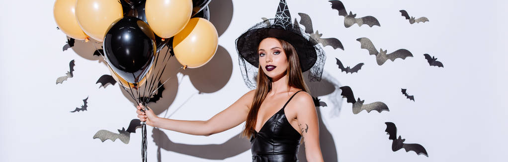 tiro panorâmico de menina em traje de bruxa negra Halloween segurando balões perto da parede branca com morcegos decorativos
 - Foto, Imagem
