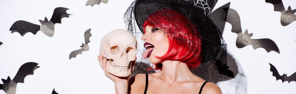 panoramic shot of girl in black witch Halloween costume with red hair licking skull near white wall with decorative bats - Photo, Image