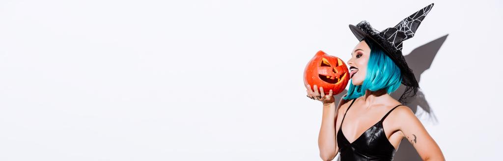 panoramic shot of sexy girl in black witch Halloween costume with blue hair licking spooky carved pumpkin on white background - Photo, Image