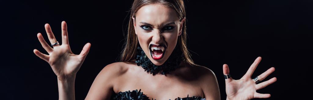 panoramiczne ujęcie przerażającej wampirzej dziewczyny z kłami w czarnej gotyckiej sukience odizolowanej na czarno - Zdjęcie, obraz