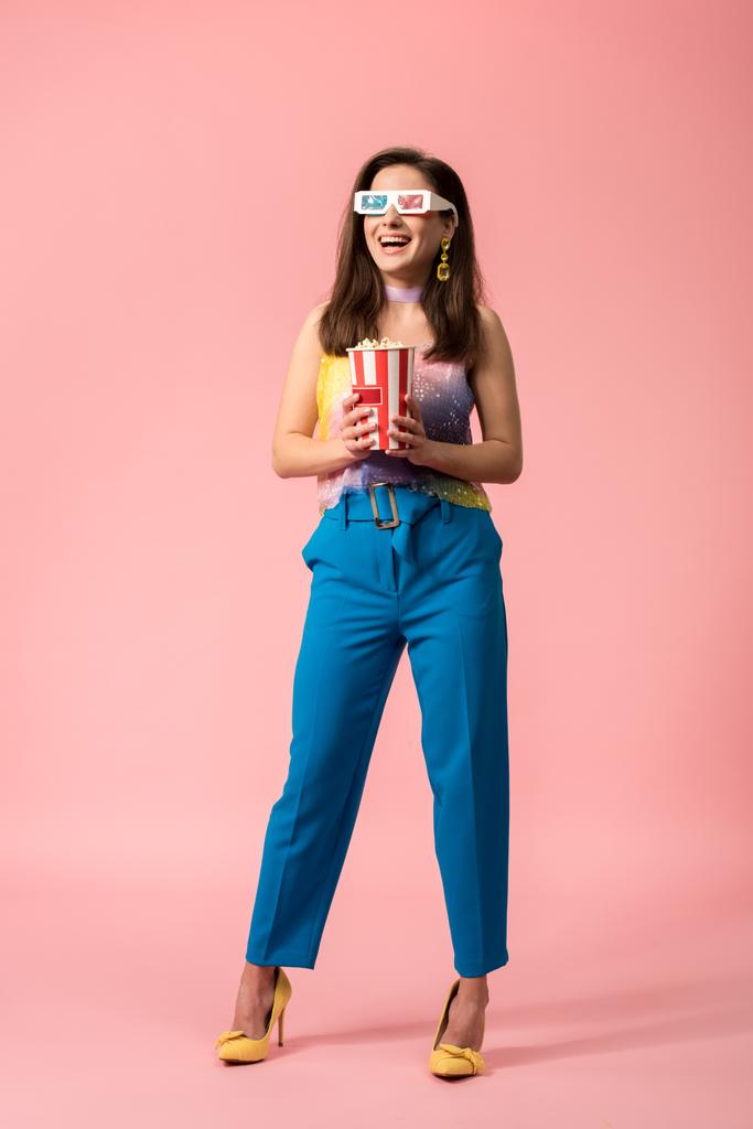 full length view of happy young stylish disco girl in 3d glasses holding striped paper bucket with popcorn on pink - Photo, Image