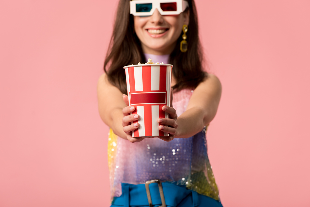 selective focus of happy young stylish disco girl in 3d glasses presenting striped paper bucket with popcorn isolated on pink - Photo, Image