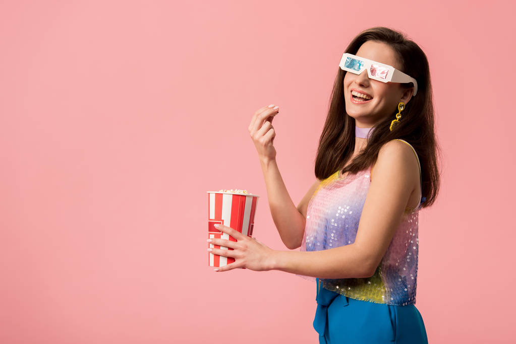 Seitenansicht von glücklichen jungen stilvollen Disco-Mädchen in 3D-Gläsern, die Popcorn isoliert auf rosa essen - Foto, Bild