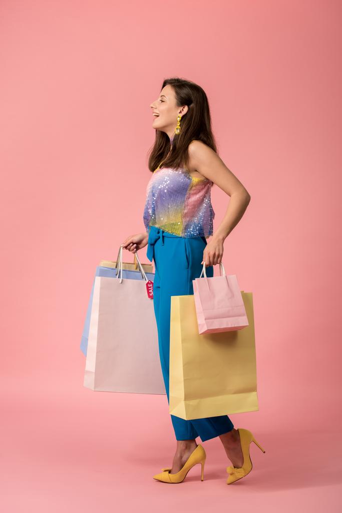 Seitenansicht des glücklichen stilvollen Disco-Mädchens, das mit Einkaufstaschen auf rosa läuft - Foto, Bild