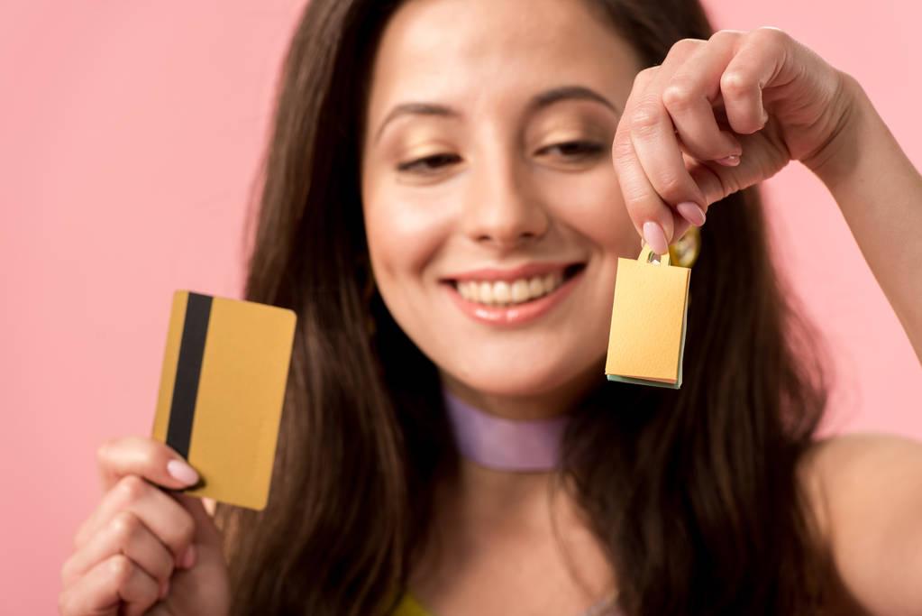 selective focus of happy stylish disco girl holding shopping bag and credit card isolated on pink - 写真・画像