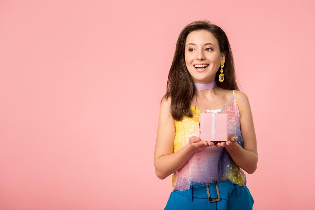 happy young stylish disco girl holding gift box and looking away isolated on pink - 写真・画像