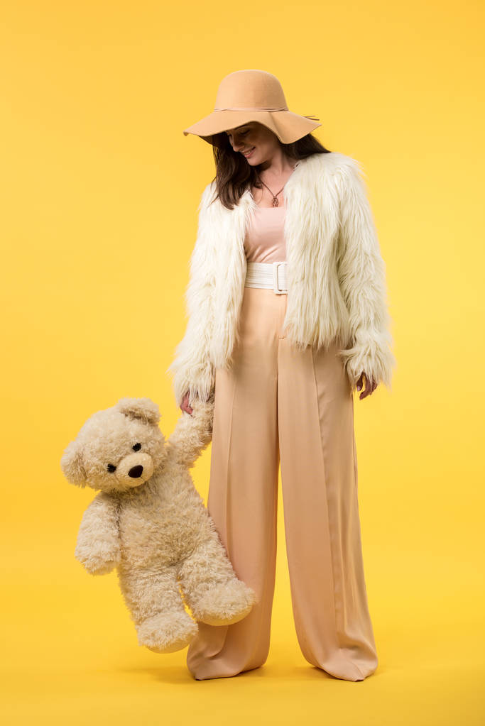 happy elegant girl in faux fur jacket and hat looking at teddy bear on yellow - Photo, Image