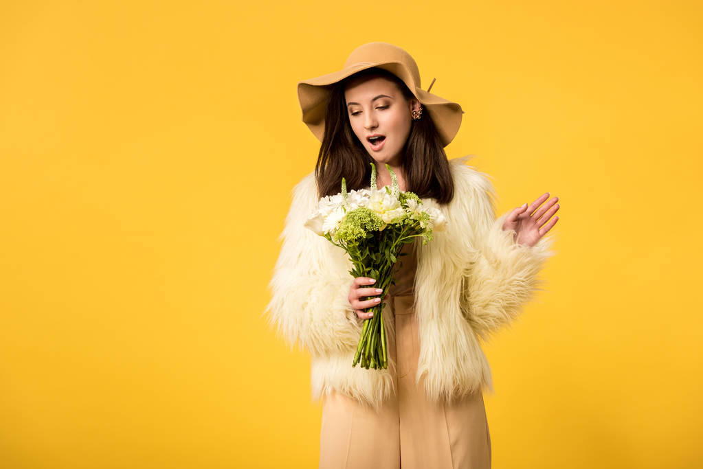 šokované elegantní dívka ve falešné kožešinové bundě a klobouk při pohledu na kytici izolované na žluté - Fotografie, Obrázek