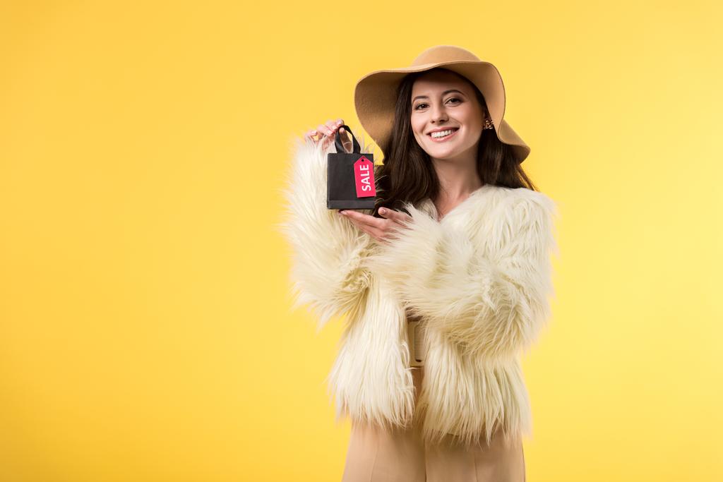 χαμογελαστό κομψό κορίτσι σε faux γούνα σακάκι και καπέλο κρατώντας τσάντα για ψώνια με την πώληση επιστολόχαρτα απομονώνονται σε κίτρινο - Φωτογραφία, εικόνα