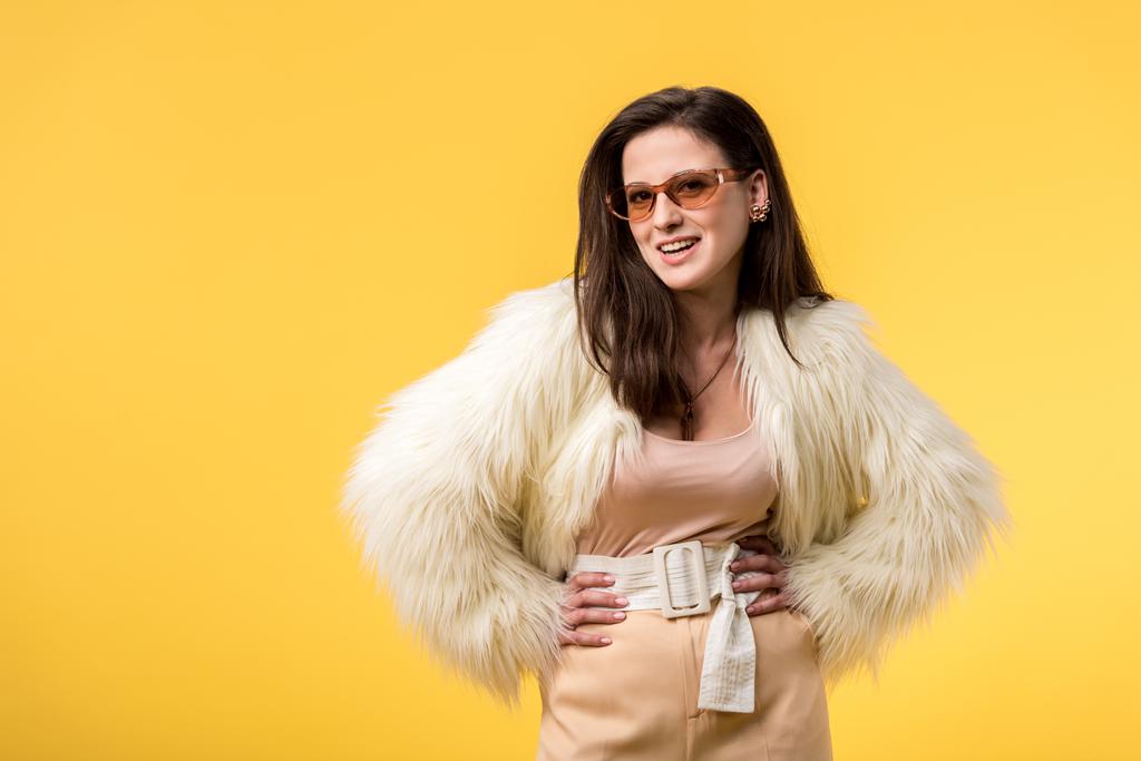 smiling party girl in faux fur jacket and sunglasses with hands on hips isolated on yellow - Photo, Image