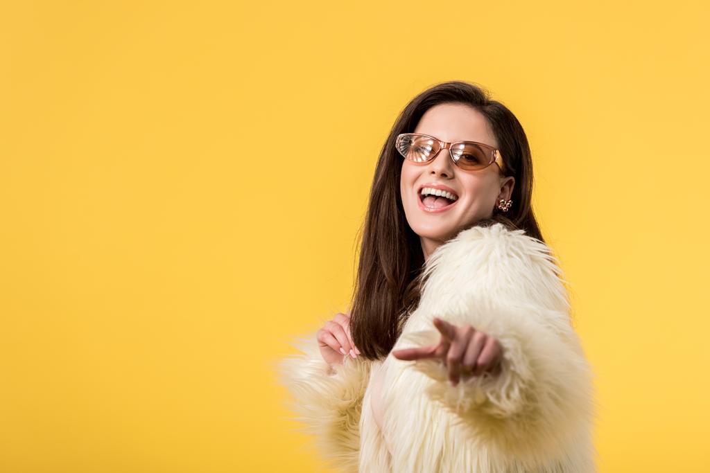 izgatott party lány faux szőrme kabát és napszemüveg mutató ujjal kamera elszigetelt sárga - Fotó, kép