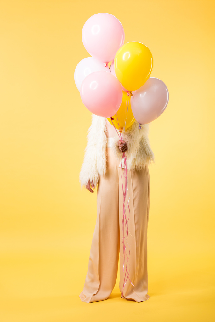 party girl in faux fur jacket holding balloons in front of face on yellow background - 写真・画像