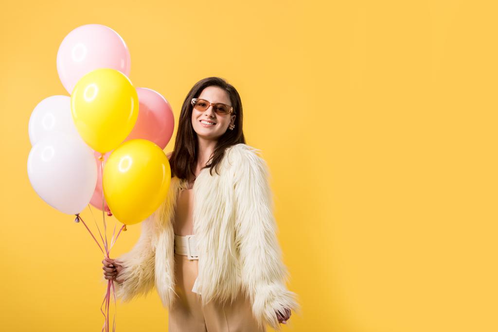 χαμογελαστό κορίτσι κόμμα σε faux γούνα σακάκι και γυαλιά ηλίου κρατώντας μπαλόνια που απομονώνονται σε κίτρινο - Φωτογραφία, εικόνα