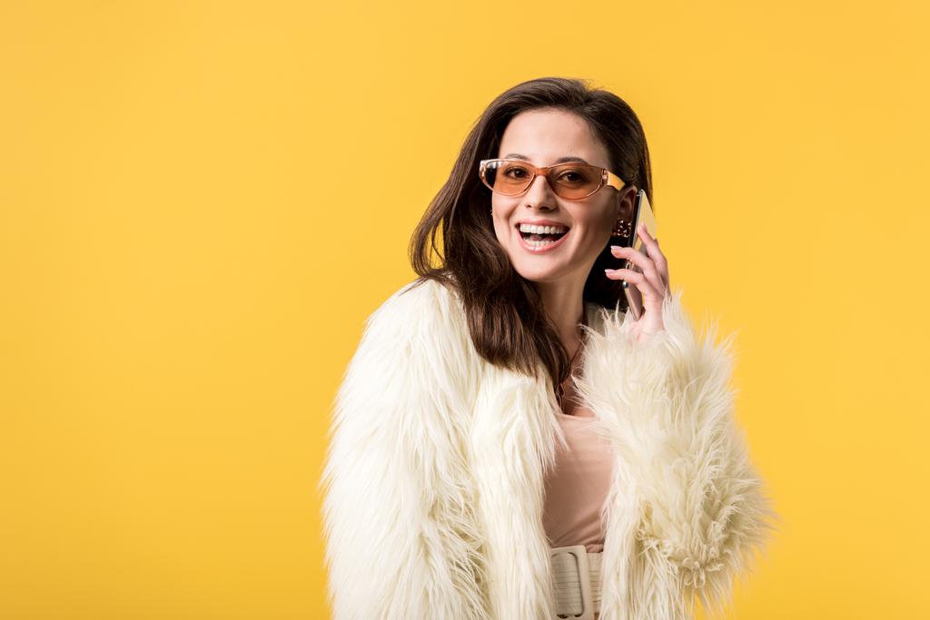 chica fiesta feliz en chaqueta de piel sintética y gafas de sol hablando en el teléfono inteligente aislado en amarillo
 - Foto, Imagen