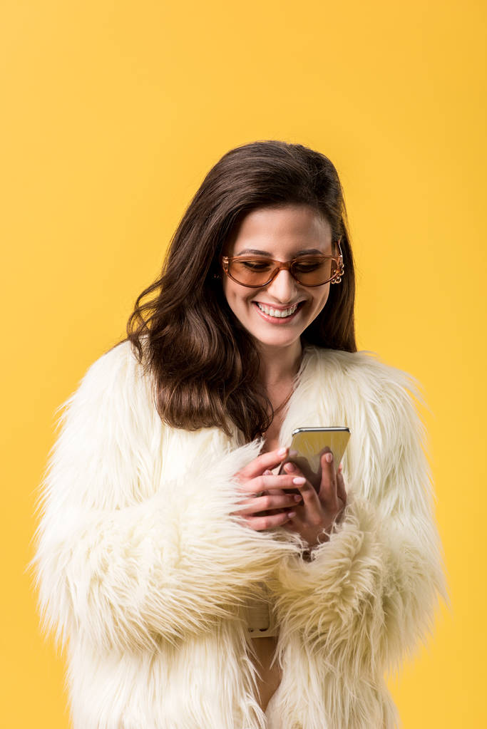 χαρούμενο κορίτσι κόμμα σε faux γούνα σακάκι και γυαλιά ηλίου χρησιμοποιώντας smartphone απομονωμένο σε κίτρινο - Φωτογραφία, εικόνα