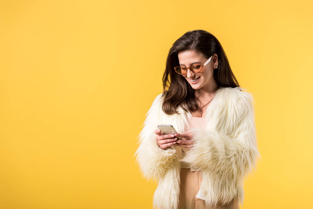 happy party girl in faux fur jacket and sunglasses using smartphone isolated on yellow - 写真・画像