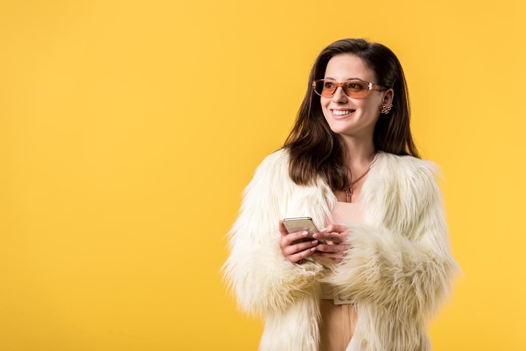 Sahte kürklü ve güneş gözlüklü mutlu parti kızı sarıda izole edilmiş akıllı telefon tutuyor. - Fotoğraf, Görsel