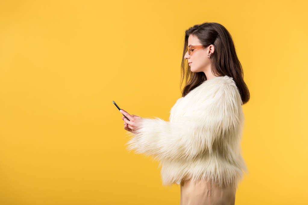 Seitenansicht des Partygirls in Kunstpelzjacke und Sonnenbrille, das Smartphone isoliert auf Gelb haltend - Foto, Bild