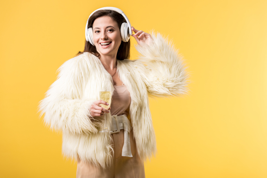 χαμογελαστό κορίτσι σε faux γούνα σακάκι ακούγοντας μουσική στα ακουστικά και κρατώντας ένα ποτήρι σαμπάνια που απομονώνονται σε κίτρινο - Φωτογραφία, εικόνα