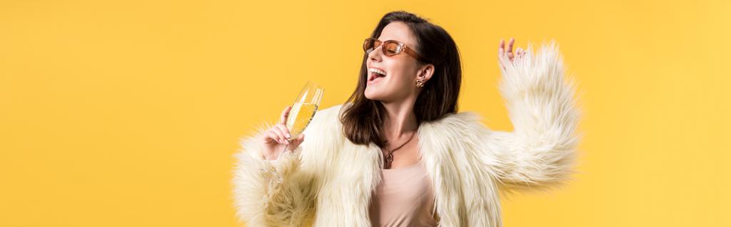 panoramic shot of happy party girl in faux fur jacket and sunglasses dancing with champagne isolated on yellow - 写真・画像
