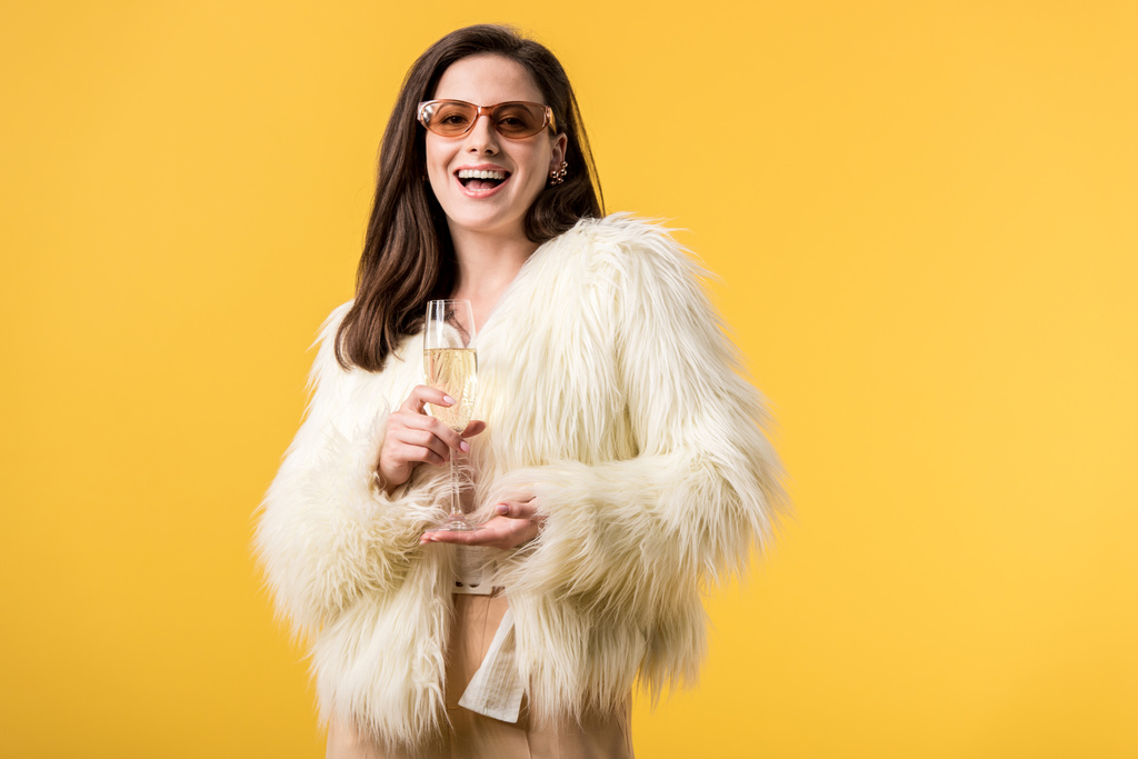 excited party girl in faux fur jacket and sunglasses with champagne isolated on yellow - 写真・画像