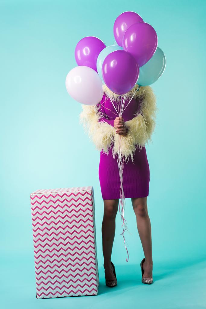chica de fiesta en vestido púrpura con plumas sosteniendo globos en frente de la cara cerca de la caja de regalo enorme en turquesa
  - Foto, Imagen