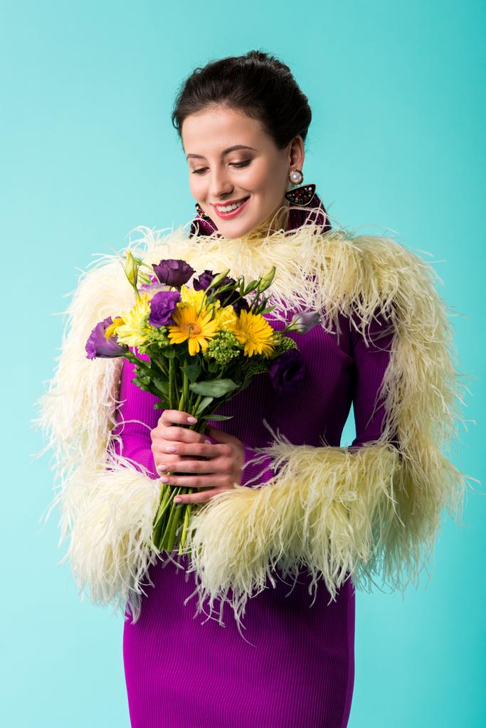 χαμογελαστό κορίτσι κόμμα σε μωβ φόρεμα με φτερά κρατώντας μπουκέτο λουλούδια που απομονώνονται σε τυρκουάζ  - Φωτογραφία, εικόνα