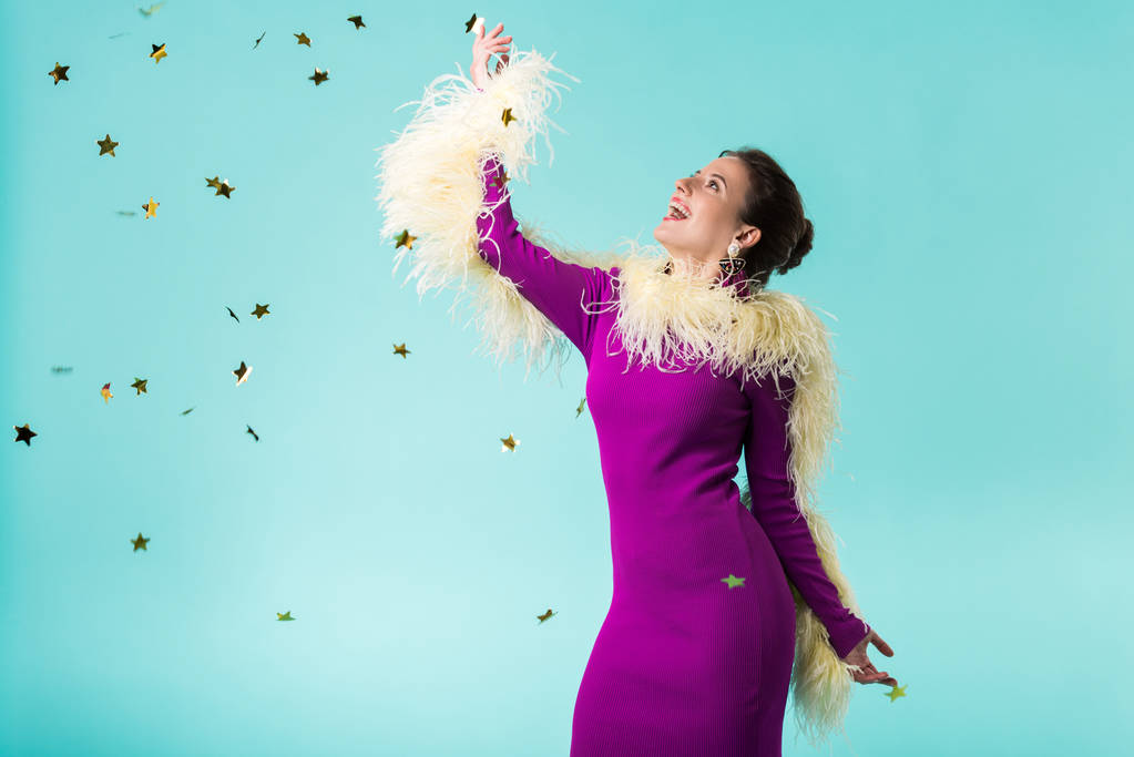 happy party girl in purple dress with feathers dancing under falling confetti isolated on turquoise  - Photo, Image