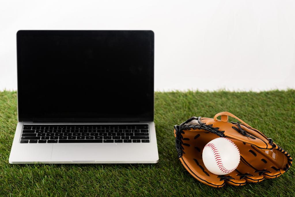 rękawica baseballowa i piłka w pobliżu laptopa z pustym ekranem na zielonej trawie izolowane na białym, koncepcja zakładów sportowych - Zdjęcie, obraz