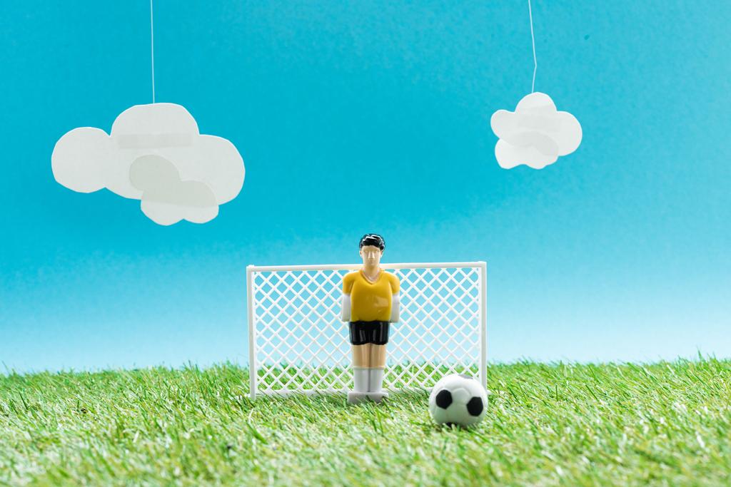 voetbalveld met speelgoedkeeper in de buurt van miniatuur voetbal op blauwe achtergrond met wolken, sportweddenschappen concept - Foto, afbeelding