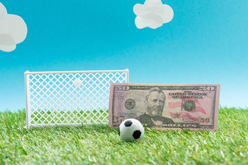игрушечный футбольный мяч и ворота возле долларовой купюры на синем фоне с облаками, концепция ставок на спорт
 - Фото, изображение