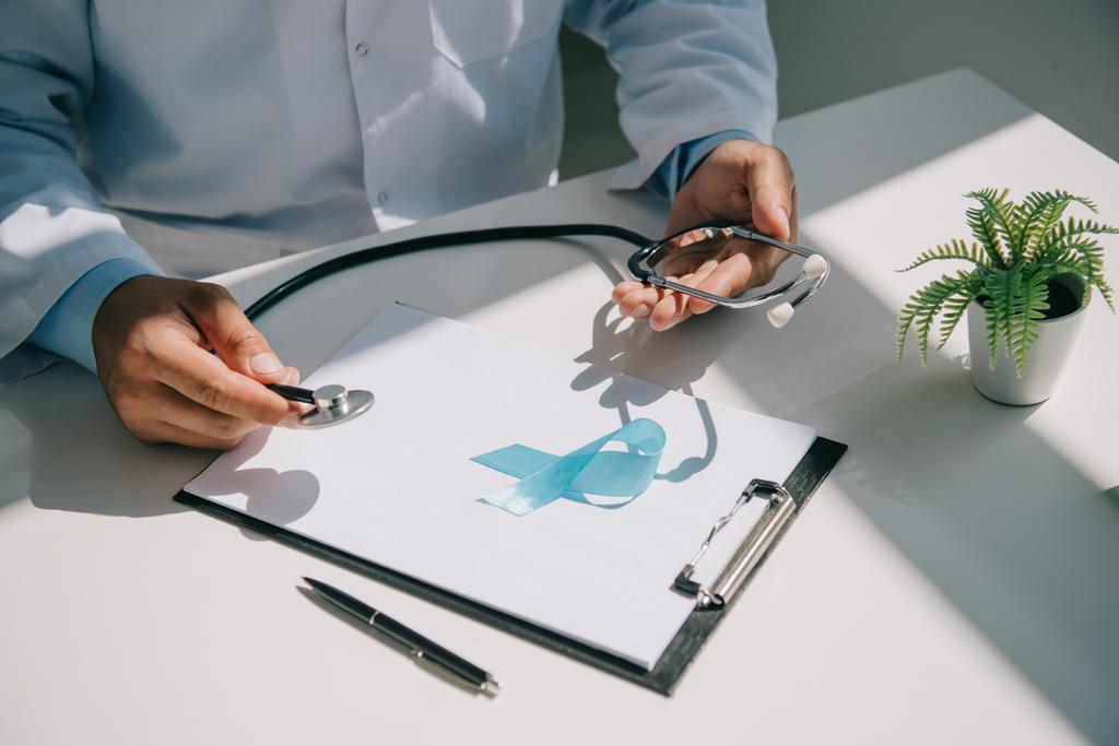 gedeeltelijke weergave van arts die stethoscoop in de buurt van blauw bewustmakingslint op klembord met blanco papier - Foto, afbeelding