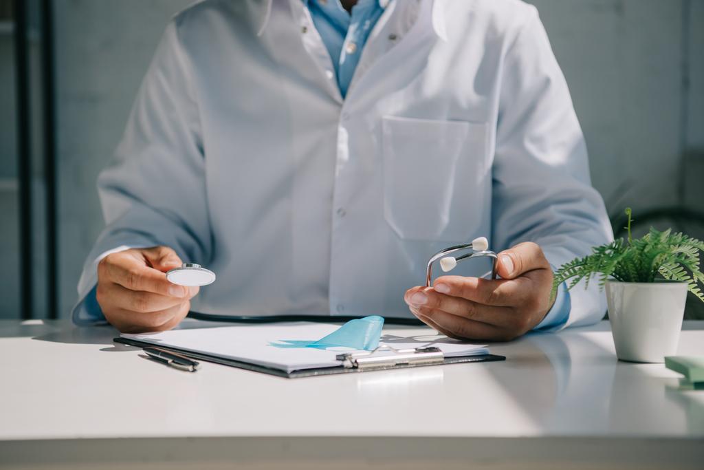 Masada oturan ve steteskopu mavi farkındalık kurdelesinin yanında tutan doktorların kesikli görüntüsü. - Fotoğraf, Görsel
