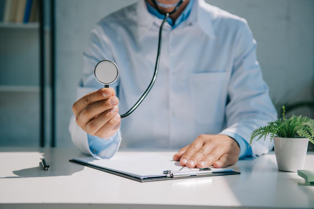 oříznutý pohled na lékaře držícího stetoskop při sezení za stolem - Fotografie, Obrázek