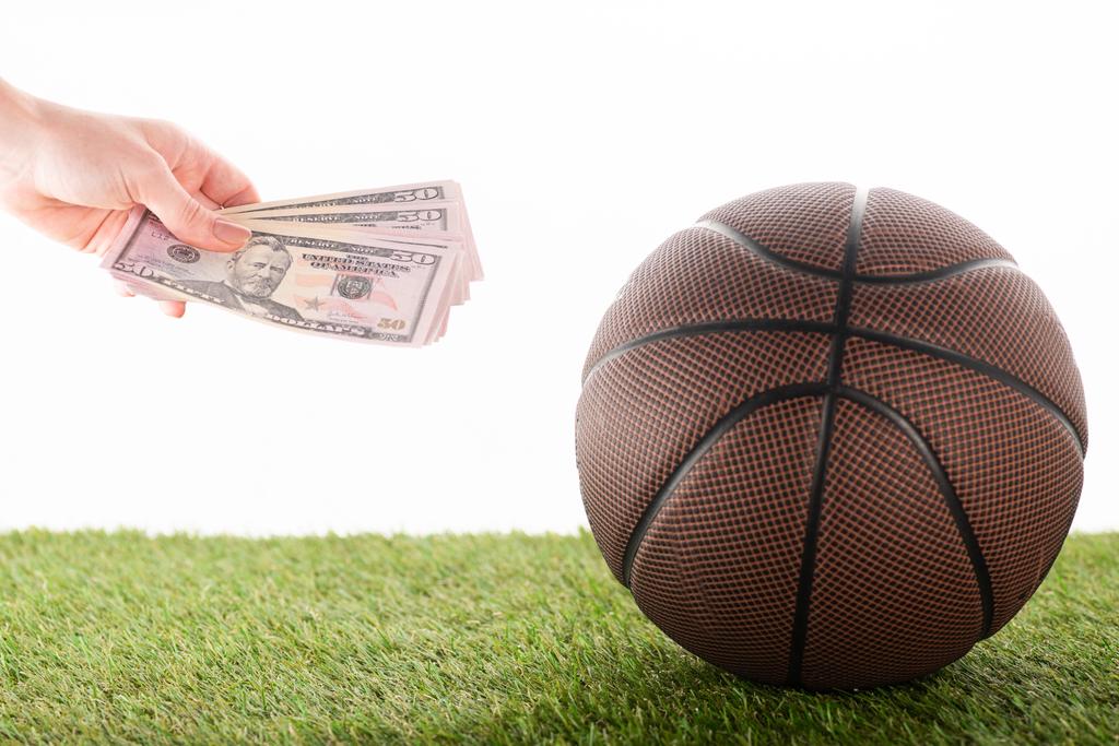 przycięty widok kobiecej ręki z banknotami dolara w pobliżu piłki do koszykówki na zielonej trawie odizolowany na białym, koncepcja zakładów sportowych - Zdjęcie, obraz
