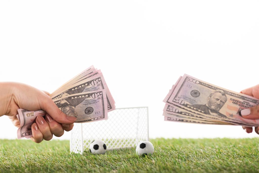 bijgesneden beeld van vrouwelijke handen met dollarbiljetten in de buurt van speelgoed voetbal ballen en poorten op groen gras geïsoleerd op wit, sportweddenschappen concept - Foto, afbeelding