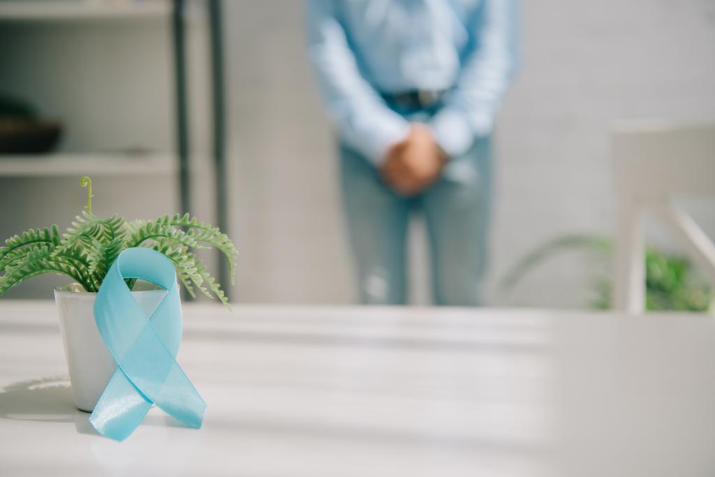 selektiver Fokus des blauen Bewusstseinsbands in der Nähe von Blumentopf und Mann, der an der Wand steht und geballte Hände in der Nähe der Leistengegend hält - Foto, Bild