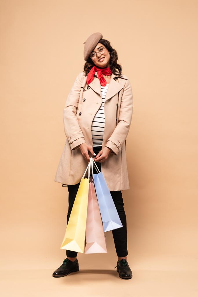 zwanger frans vrouw in jas houden boodschappentassen op beige achtergrond  - Foto, afbeelding