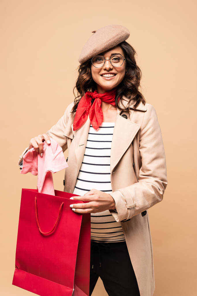 Schwangere Französin im Mantel nimmt Overalls aus Einkaufstasche auf beigem Hintergrund  - Foto, Bild